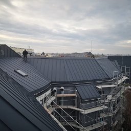 Przemysław Rejek Dekarstwo - Nowoczesny, grafitowy dach z blachy na wielospadowym budynku. Na dachu stoi dekarz. Widoczne rusztowania i okno dachowe. Pochmurne niebo w tle.