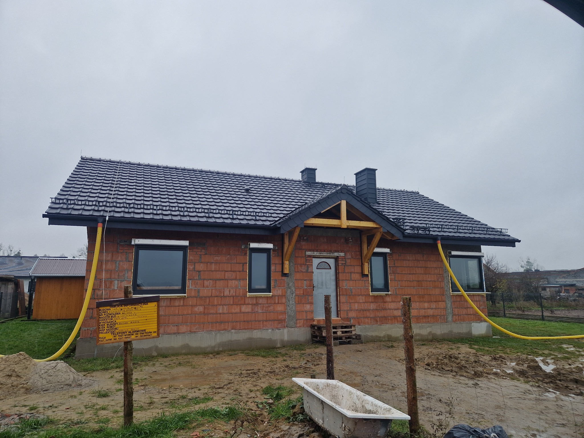 Dom w trakcie budowy z nowym, ciemnoszarym dachem z dachówki. Widoczne okna, surowa elewacja z cegły, drewniane elementy konstrukcyjne nad wejściem i żółty wąż na elewacji.