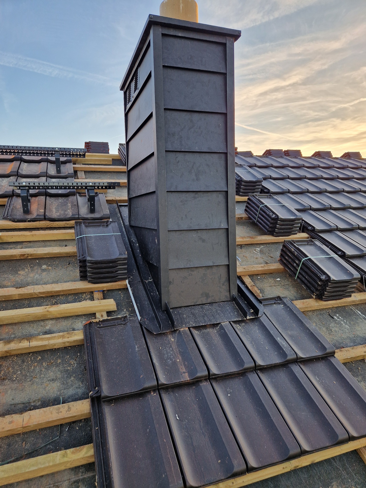 Montaż ciemnej dachówki ceramicznej wokół komina na drewnianej konstrukcji dachu. Częściowo ułożone dachówki i paczki z nowymi. Błękitne niebo w tle.