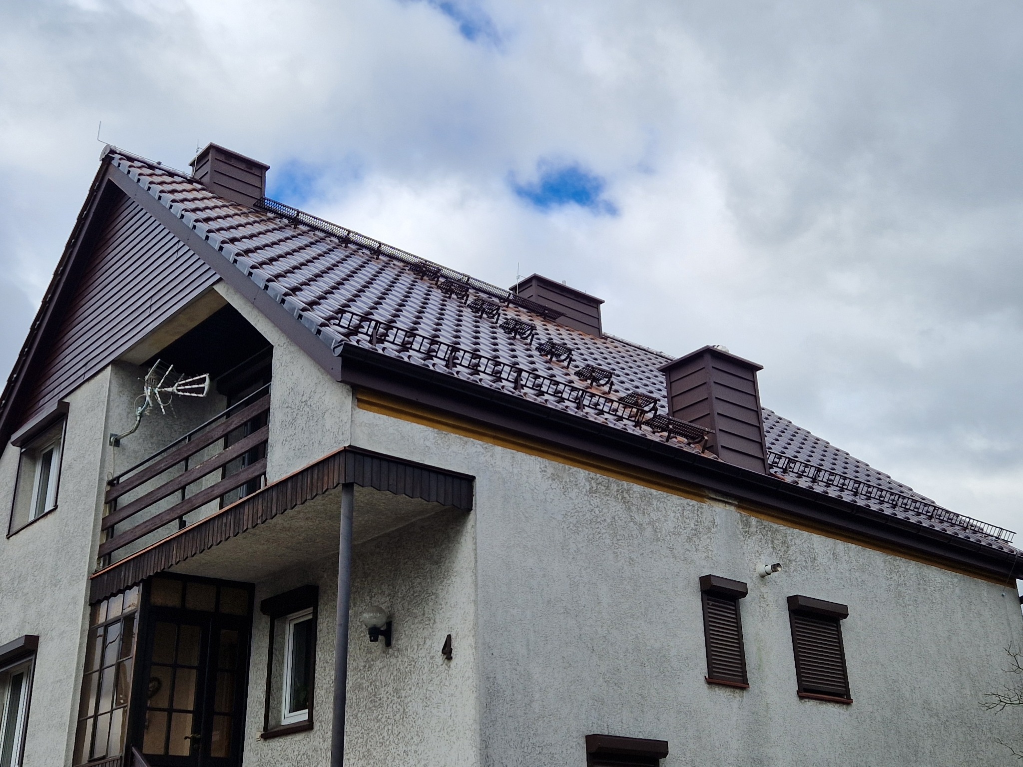 Dwupiętrowy dom z brązowym dachem z dachówki ceramicznej, brązowymi rynnami i kominami. Widoczne zabezpieczenia przeciwśniegowe na dachu. Pochmurne niebo w tle.