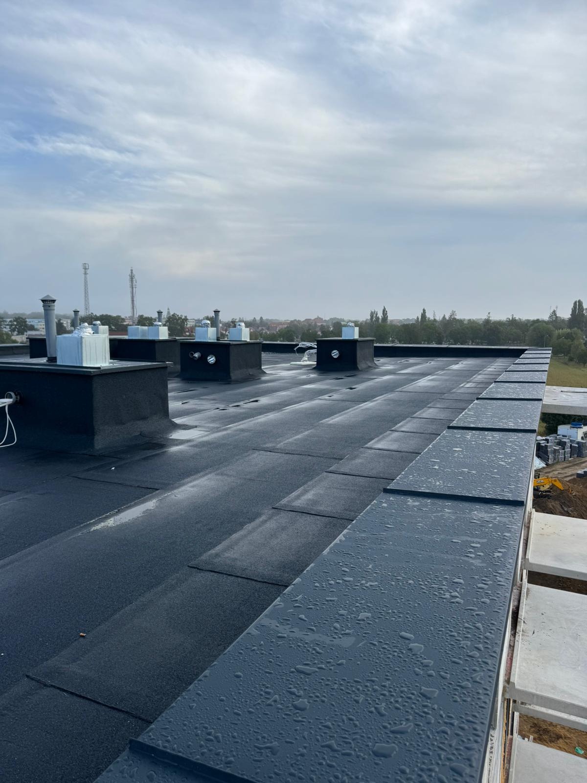 Nowy, czarny dach z papy bitumicznej z kominkami wentylacyjnymi i obróbkami blacharskimi, pokryty kroplami deszczu. W tle widoczny krajobraz miejski i budowa.