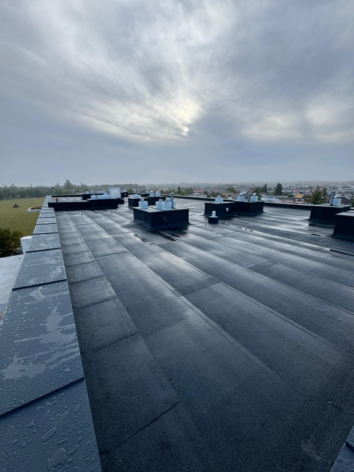 Widok z góry na płaski dach pokryty czarną papą bitumiczną z licznymi kominkami wentylacyjnymi. W tle widoczne budynki i pochmurne niebo.