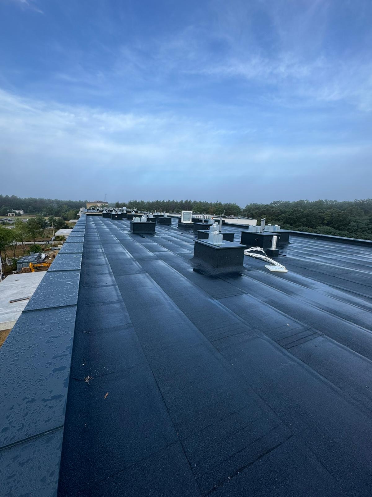 Widok z góry na płaski dach pokryty ciemną papą termozgrzewalną z kominkami wentylacyjnymi i elementami instalacji. W tle zieleń drzew i budynki w lekkiej mgle.