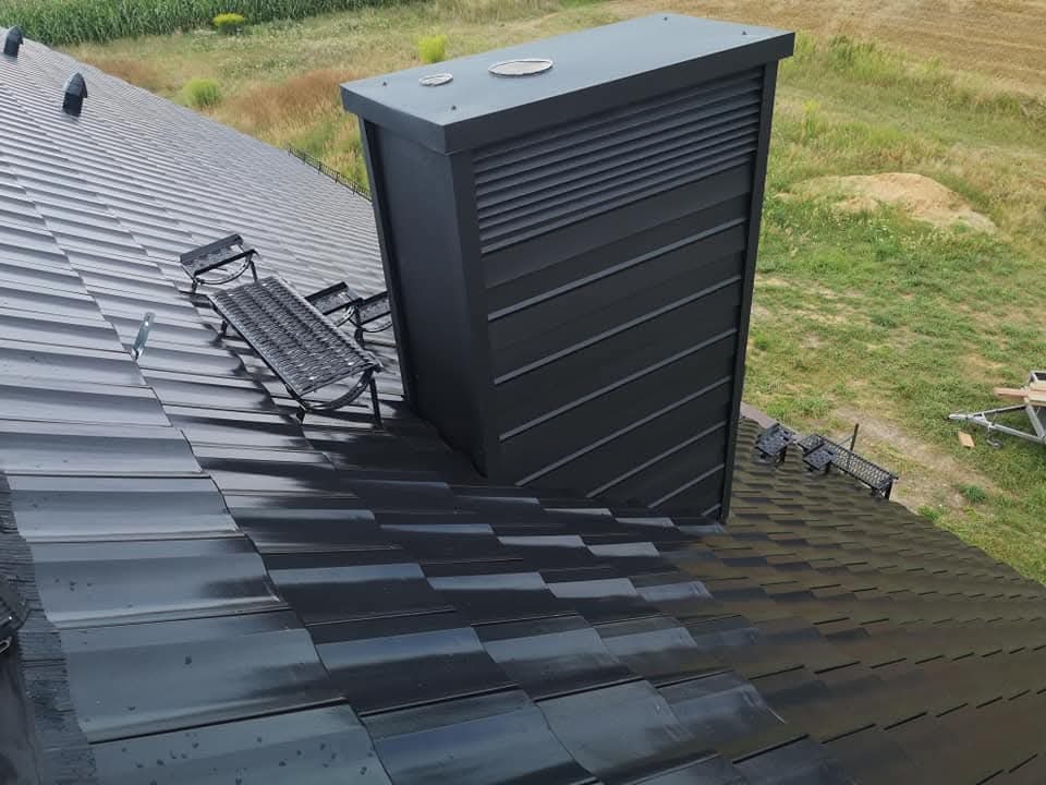 Błyszczący, grafitowy dach z blachy trapezowej z kominem wentylacyjnym i ławą kominiarską na tle zieleni. Widok z góry, ukośny kadr.