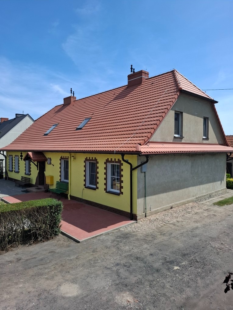 Dom z nowym, czerwonym dachem z dachówki ceramicznej, widoczny z perspektywy ulicy w słoneczny dzień. Żółta elewacja i szara ściana szczytowa.