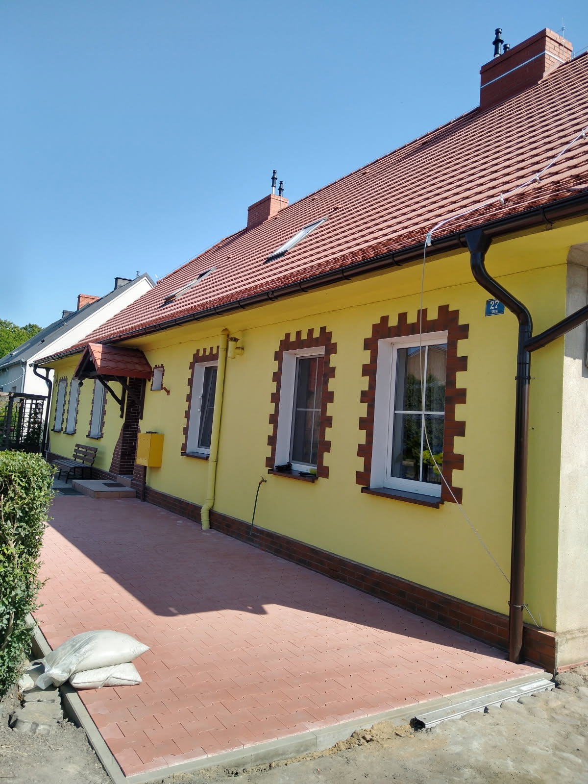 Żółty dom z czerwoną dachówką i ceglanymi detalami okiennymi. Nowa, czerwona kostka brukowa przed domem. Widoczne rynny i komin. Słoneczny dzień.