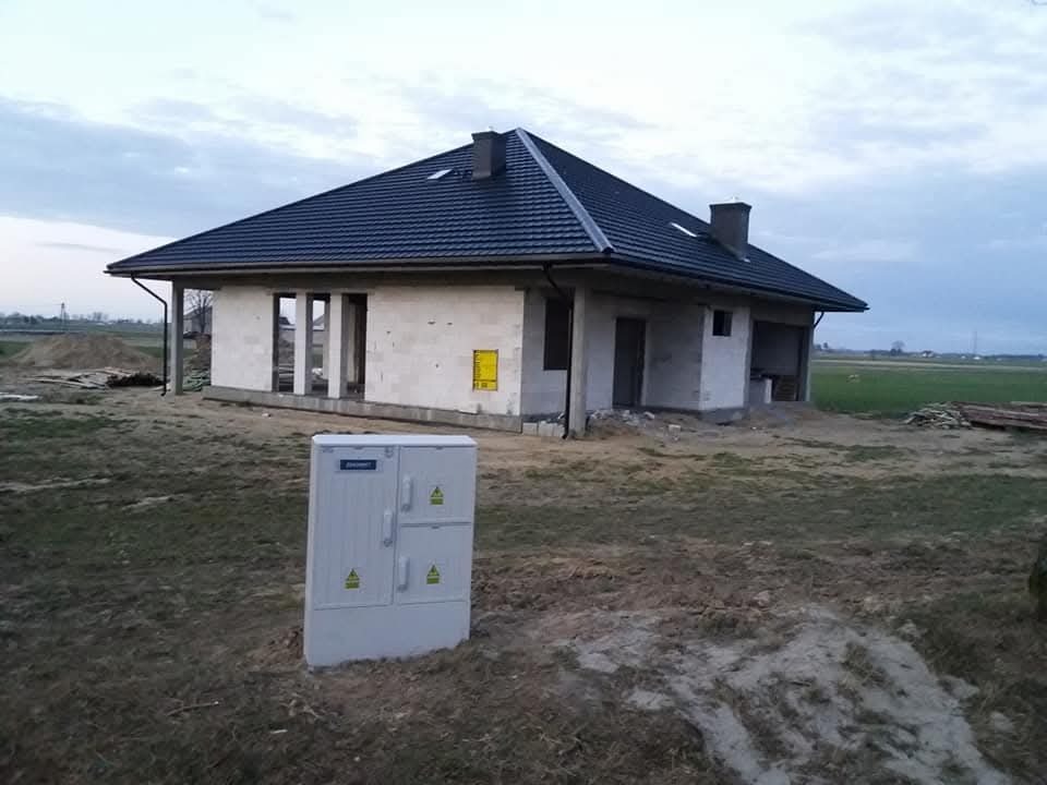 Niedokończony dom jednorodzinny z ciemnym dachem z dachówki, widoczna skrzynka elektryczna na pierwszym planie, surowy stan elewacji i otoczenia.