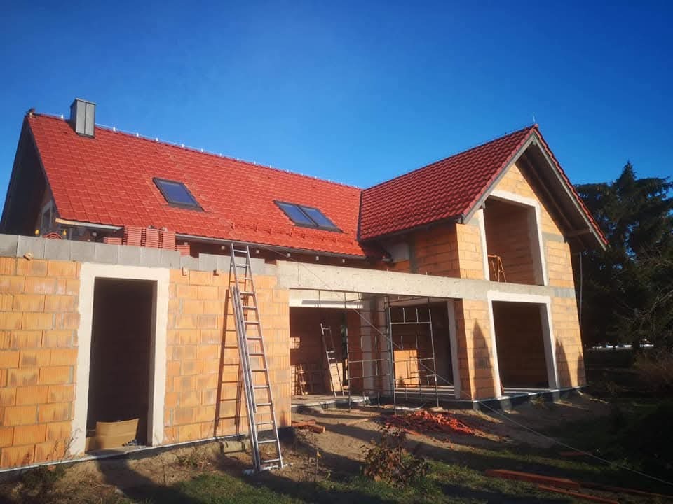 Ceglany dom w budowie z czerwonym dachem i oknami dachowymi w słoneczny dzień. Widoczna drabina i częściowo ułożone dachówki. Stan surowy otwarty.