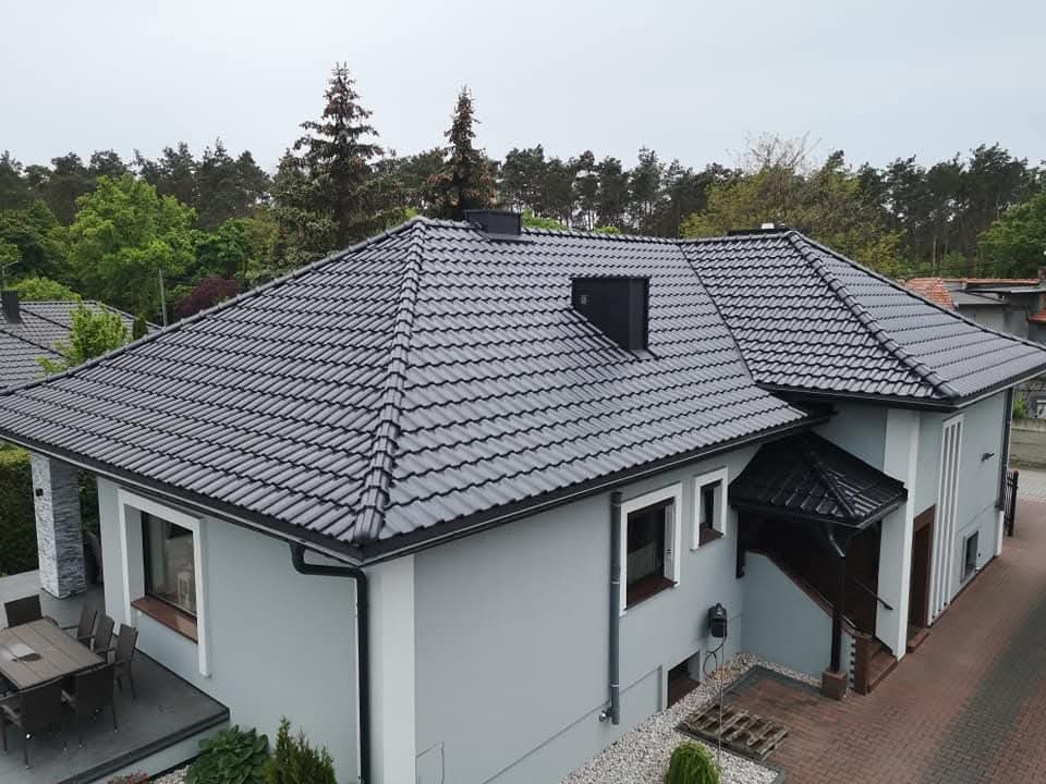 Szary dom jednorodzinny z ciemną, nową dachówką ceramiczną, widoczny z góry. Rynny w kolorze dachu, komin i obróbki blacharskie. Otoczenie: drzewa, kostka brukowa.