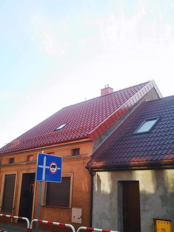 Czerwony dach z dachówki ceramicznej na dwóch budynkach, widok z dołu, znak drogowy w kadrze. Dach z oknami dachowymi, komin z cegły. Słoneczny dzień.