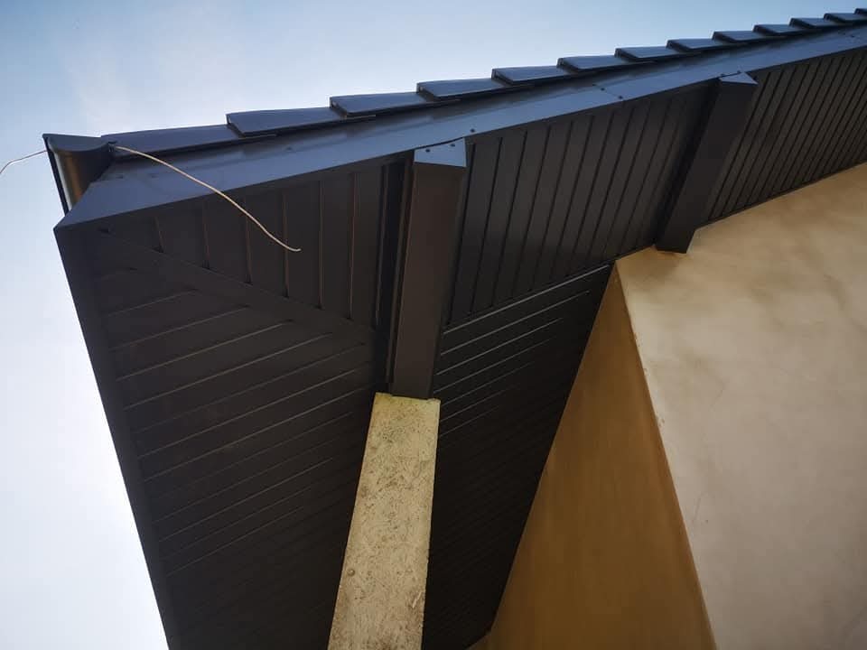 Ciemna podbitka dachowa z widoczną strukturą desek, zamontowana przy ścianie budynku o jasnej elewacji, sfotografowana pod kątem z dołu.