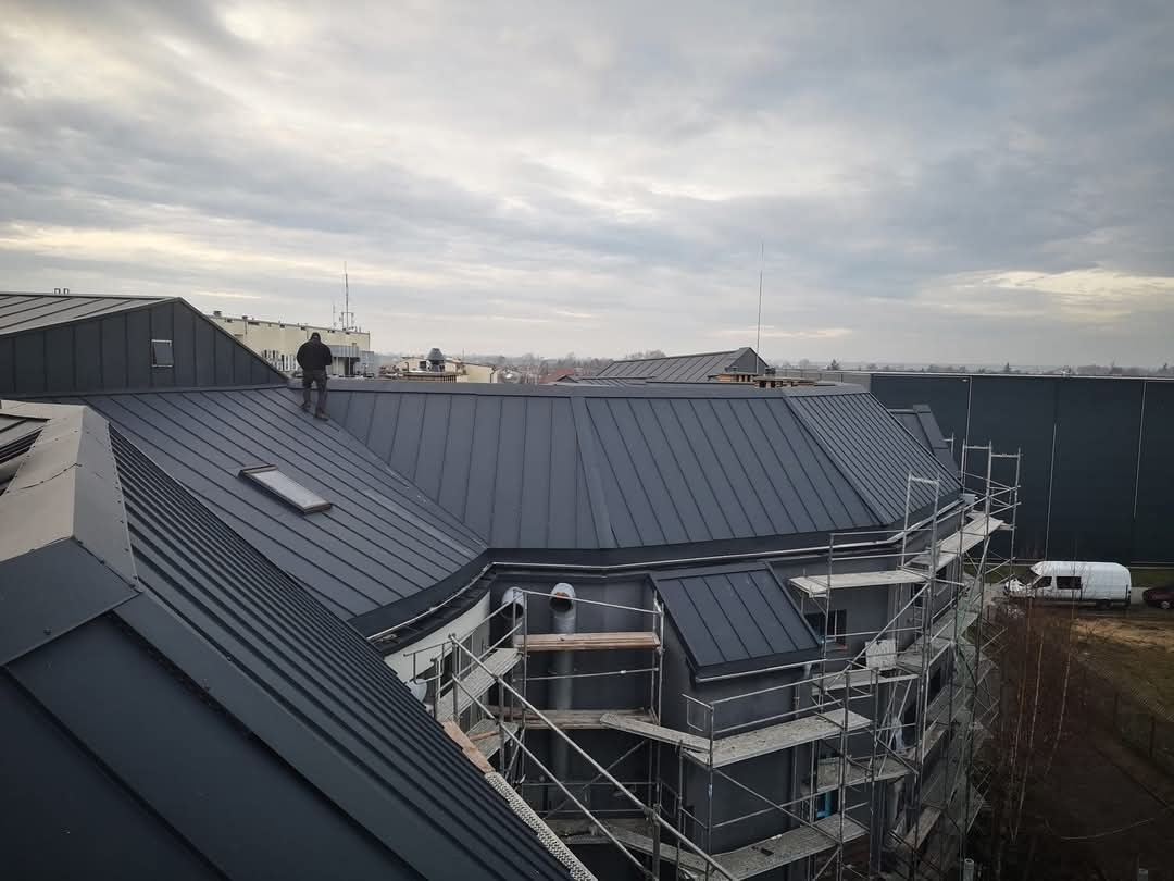 Nowoczesny, grafitowy dach z blachy na wielospadowym budynku. Na dachu stoi dekarz. Widoczne rusztowania i okno dachowe. Pochmurne niebo w tle.