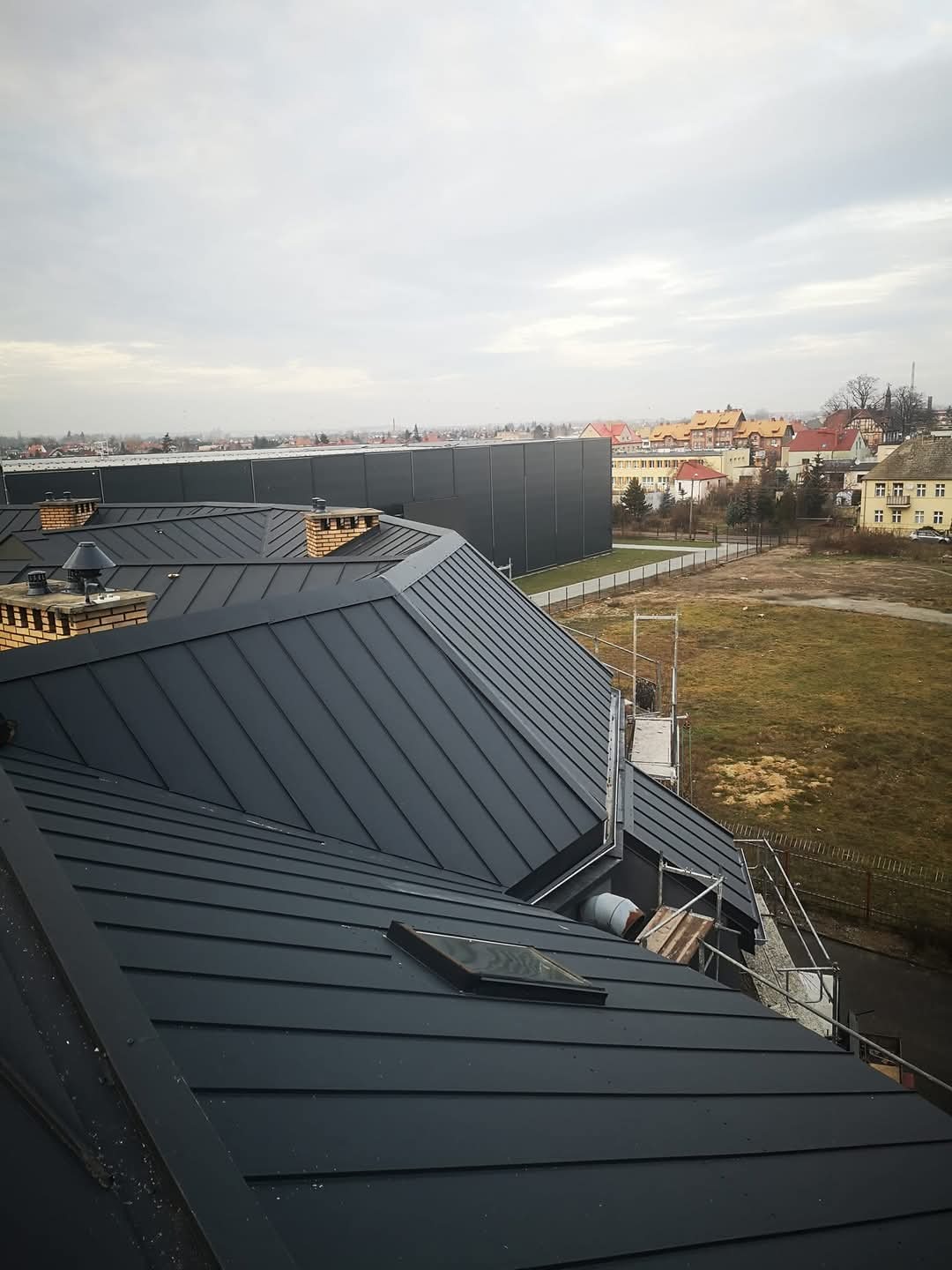 Czarny dach z blachy na budynku z oknem dachowym, widok z góry na tle zabudowań miejskich i pochmurnego nieba. Widoczne rusztowania.
