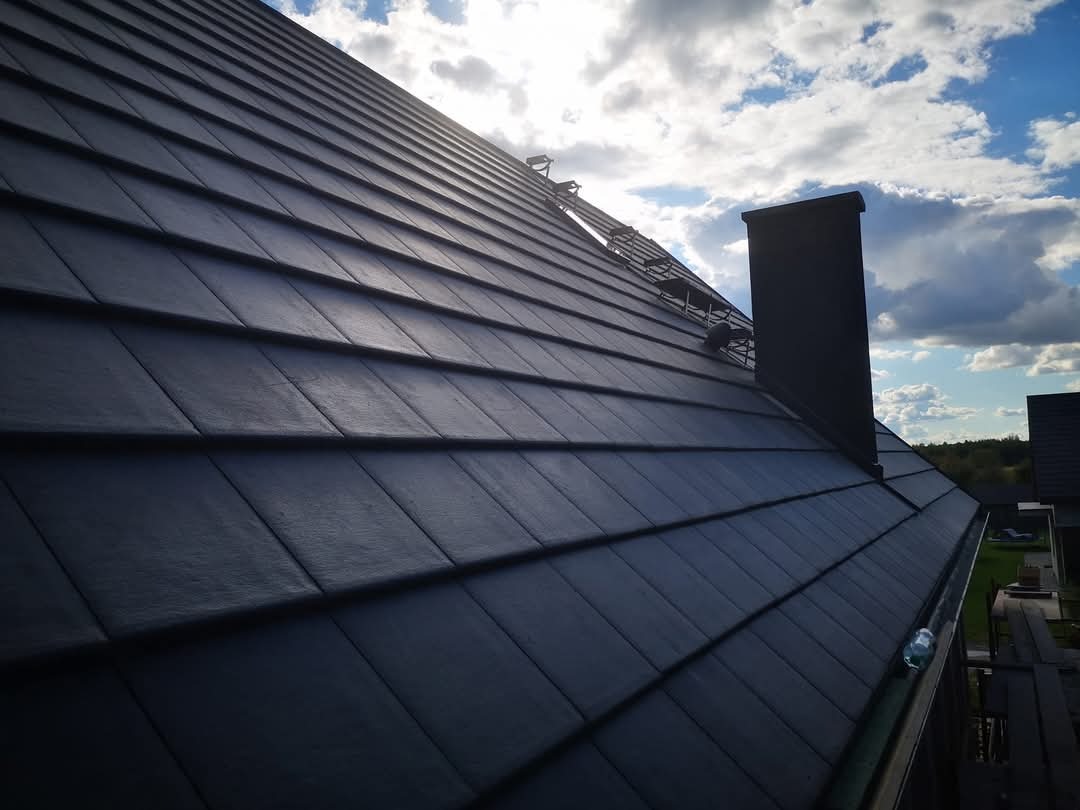 Ciemna dachówka ceramiczna na skośnym dachu domu jednorodzinnego, widok z dołu pod kątem, komin i fragment drabiny dekarskiej na tle błękitnego nieba.
