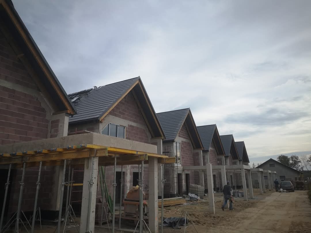 Szeregówka w trakcie budowy: wzniesione mury z cegły, częściowo pokryty dach szarą dachówką. Widoczne szalunki i podpory. Pochmurne niebo w tle.