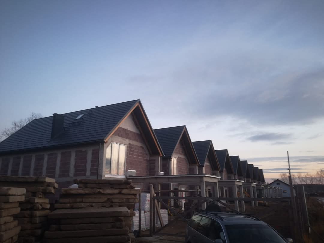 Szeregówka domów w trakcie budowy z ciemnymi dachami i widoczną konstrukcją, uchwycona o zmierzchu. Na pierwszym planie stos materiałów budowlanych.