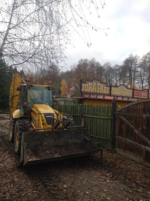 Żółta koparko-ładowarka Komatsu z łyżką pełną ziemi na tle drewnianego płotu i szyldu Jura Park. Praca w terenie, jesienna sceneria.