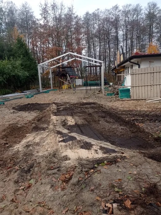 Przygotowany teren pod budowę z widoczną metalową konstrukcją szkieletową. W tle drzewa i budynek. Ślady opon na piasku.