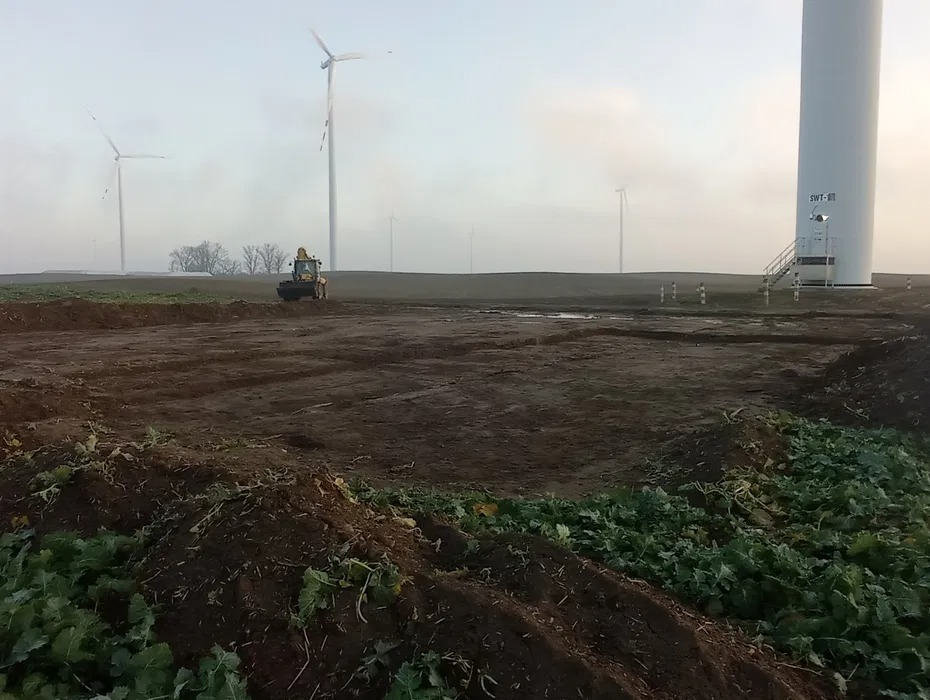 Żółta koparka na polu z turbinami w tle, przygotowany teren pod budowę. Widoczne ślady opon i wykopana ziemia. Mglista pogoda.