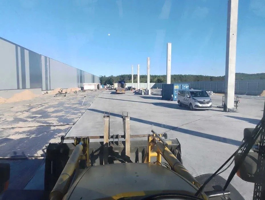Widok z kabiny traktora na plac budowy z betonową wylewką, słupami konstrukcyjnymi i zaparkowanym busem. W tle widoczny długi budynek magazynowy.
