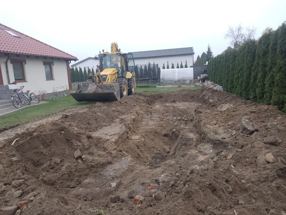 Żółta koparko-ładowarka na działce z wykopem pod fundamenty domu jednorodzinnego. W tle dom z czerwoną dachówką i wysokie tuje.