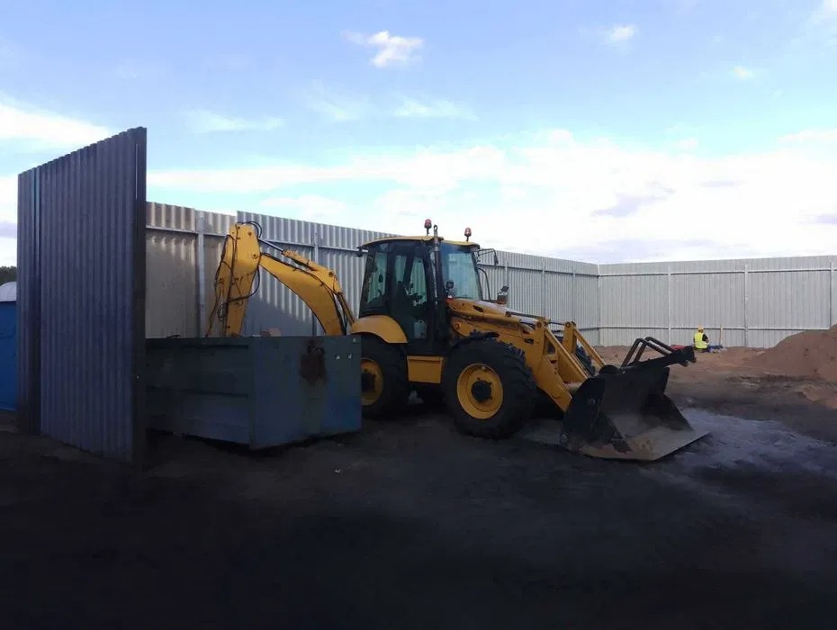 Żółta koparko-ładowarka na placu budowy, częściowo schowana za metalową konstrukcją. W tle widoczne ogrodzenie i pracownik w kamizelce odblaskowej.