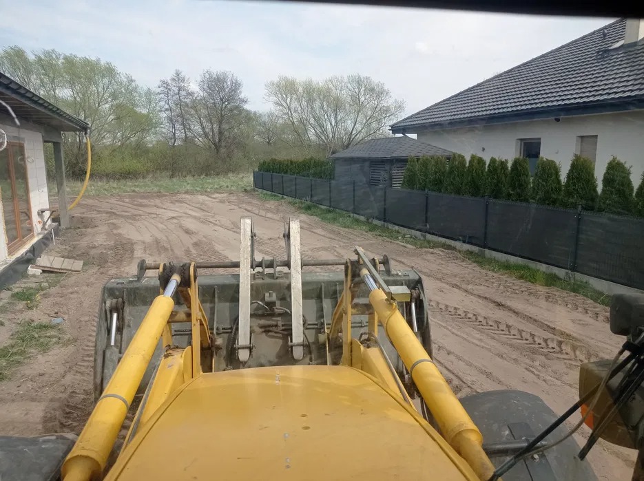 Żółta ładowarka na placu budowy, widok z kabiny operatora. W tle domy i drzewa, teren wyrównany. Ślady opon na piasku.