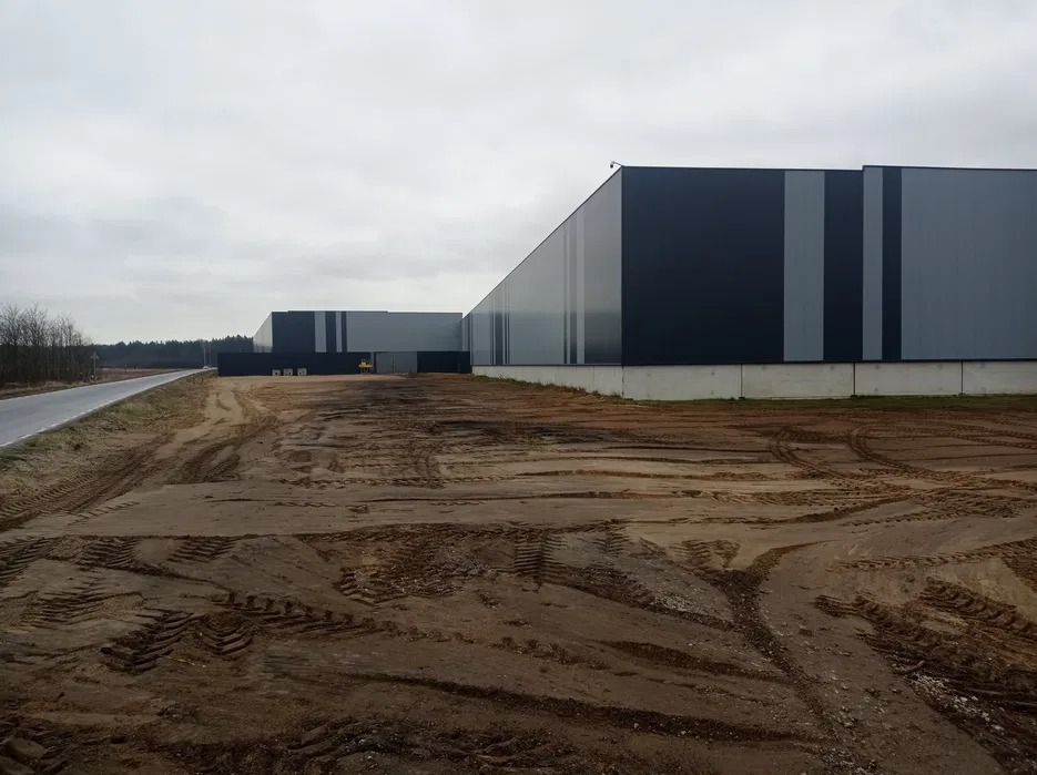 Nowoczesny budynek magazynowy z panelami w odcieniach szarości, widoczny z terenu z licznymi śladami opon, sugerującymi prace ziemne. Pochmurne niebo w tle.