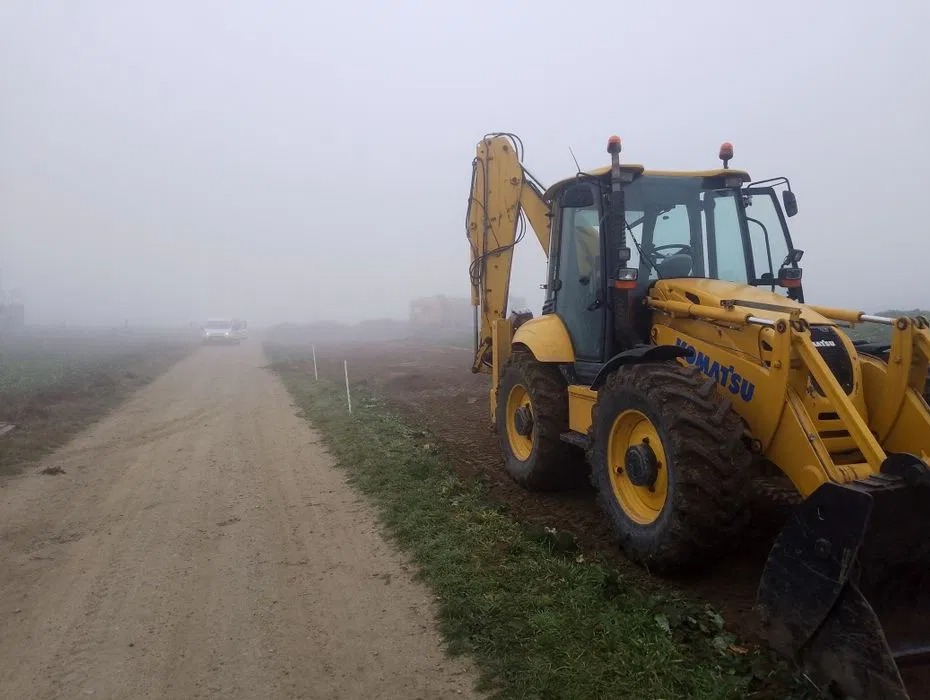 Żółta koparko-ładowarka Komatsu stoi na ziemi w mglisty dzień. W tle polna droga i niewyraźny pojazd. Widoczne ślady opon i świeżo naruszona ziemia.