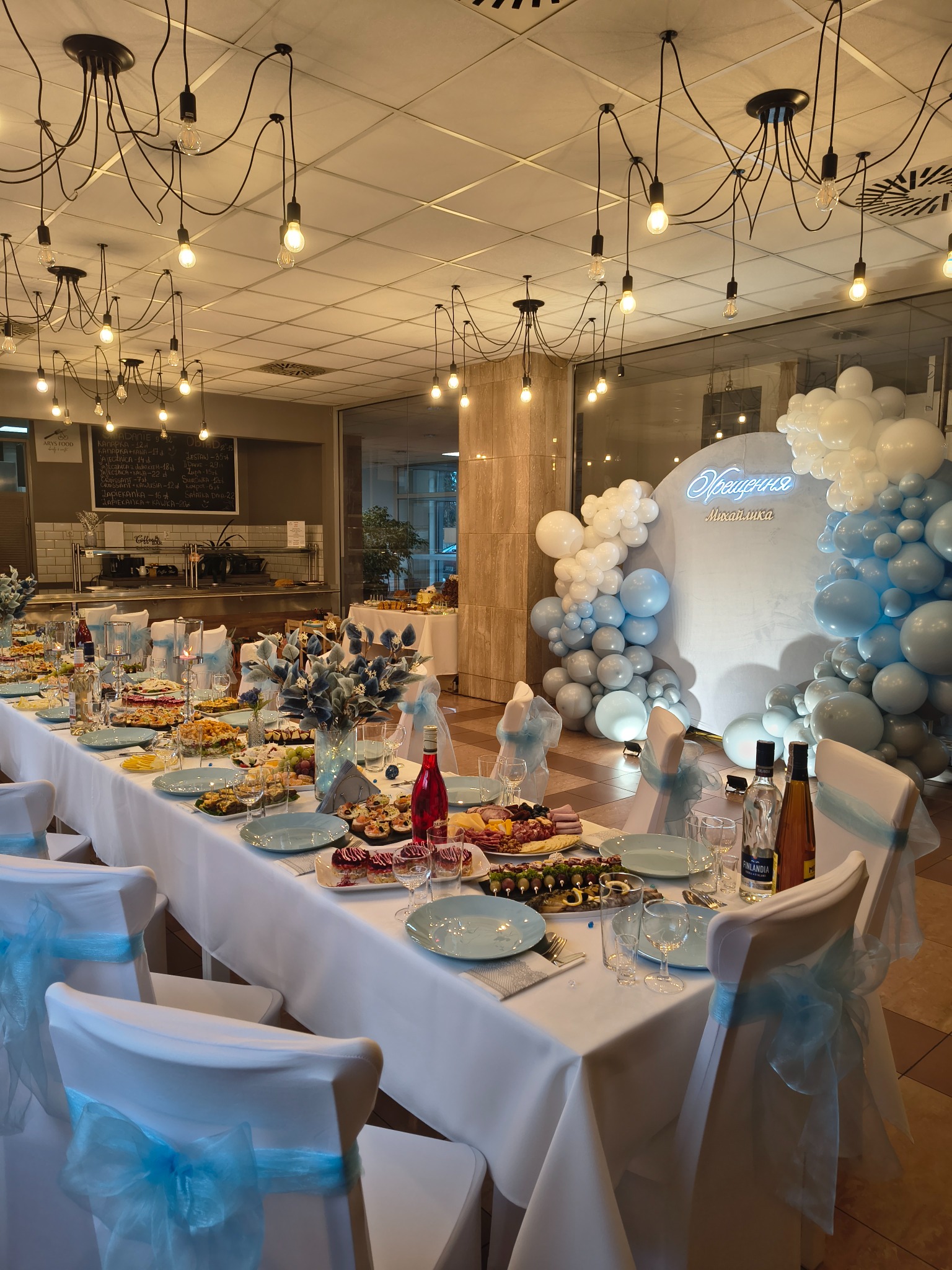 Uroczysty stół z dekoracją balonową w odcieniach błękitu i bieli, zastawiony potrawami i napojami. Krzesła z białymi pokrowcami i błękitnymi kokardami. Oświetlenie sufitowe z żarówkami.