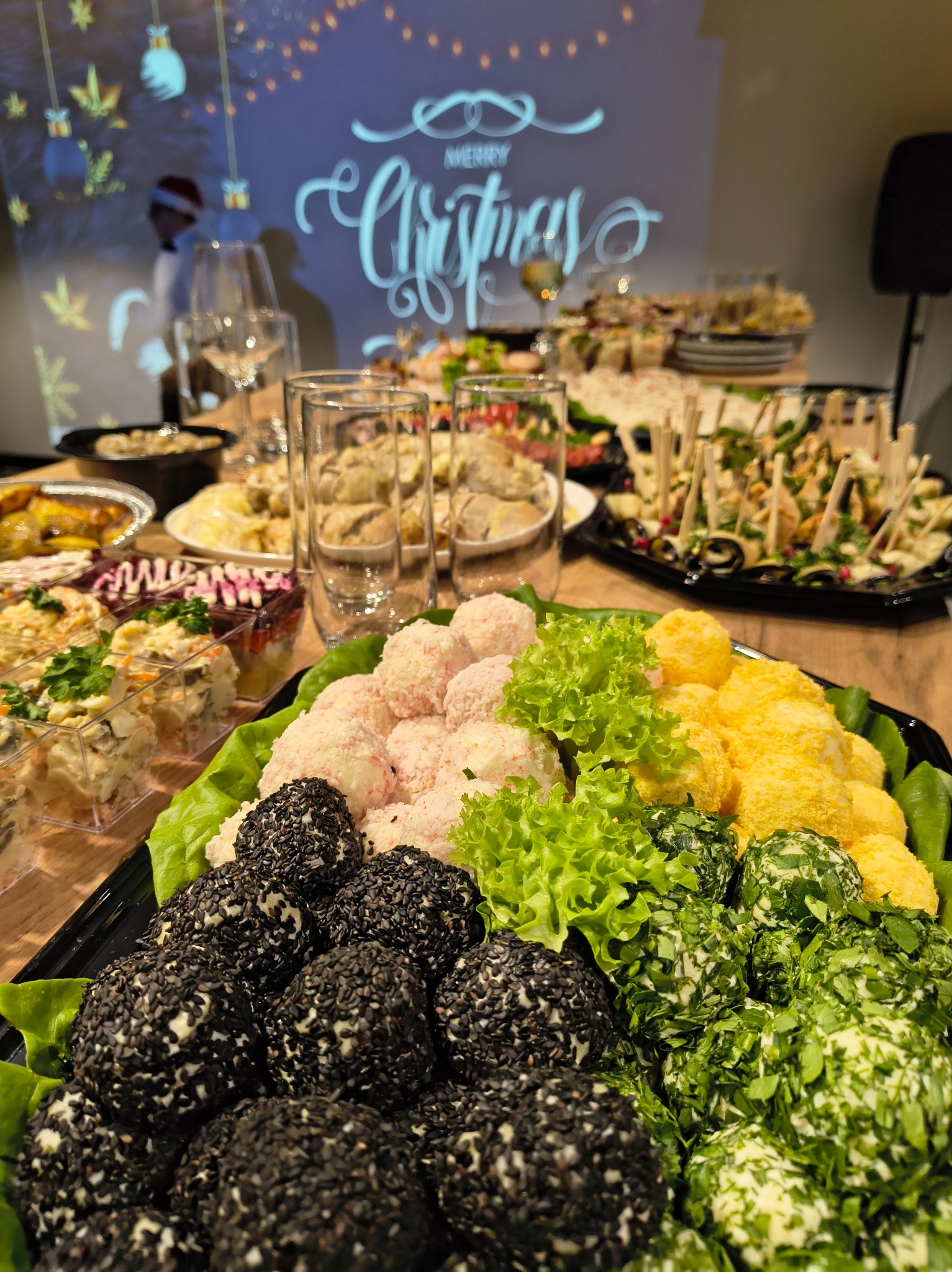 Świąteczny stół cateringowy: kolorowe przekąski na paterach, w tle dekoracja 'Merry Christmas' i kelner w czapce Mikołaja. Różnorodność smaków i świąteczny klimat.