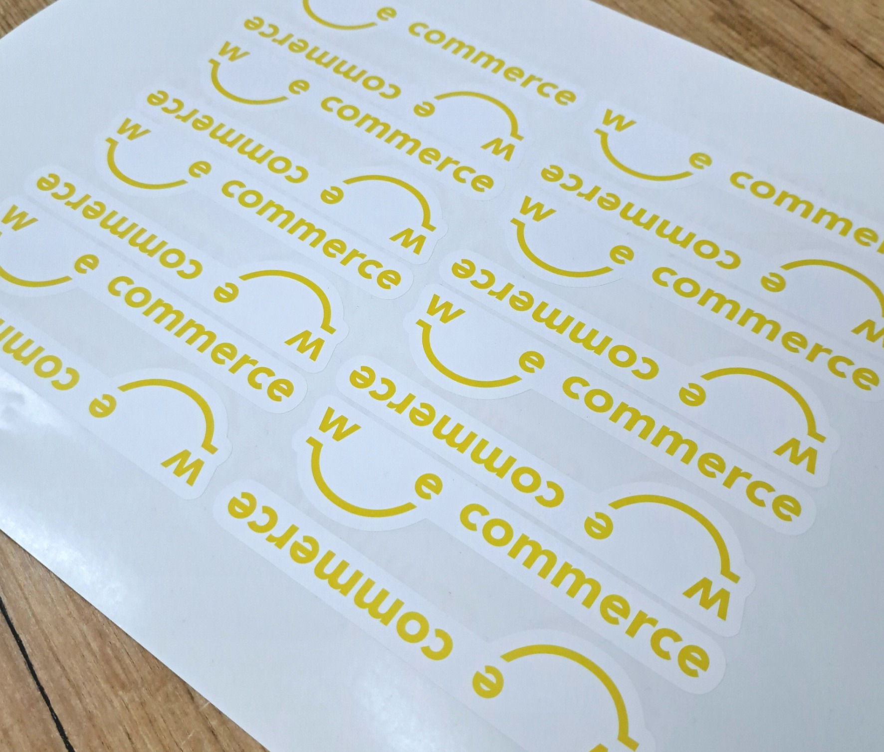Arkusz z naklejkami w kolorze żółtym z napisem 'e commerce' i symbolem uśmiechu z literą 'W' i 'M' na białym tle, leżący na drewnianej powierzchni.