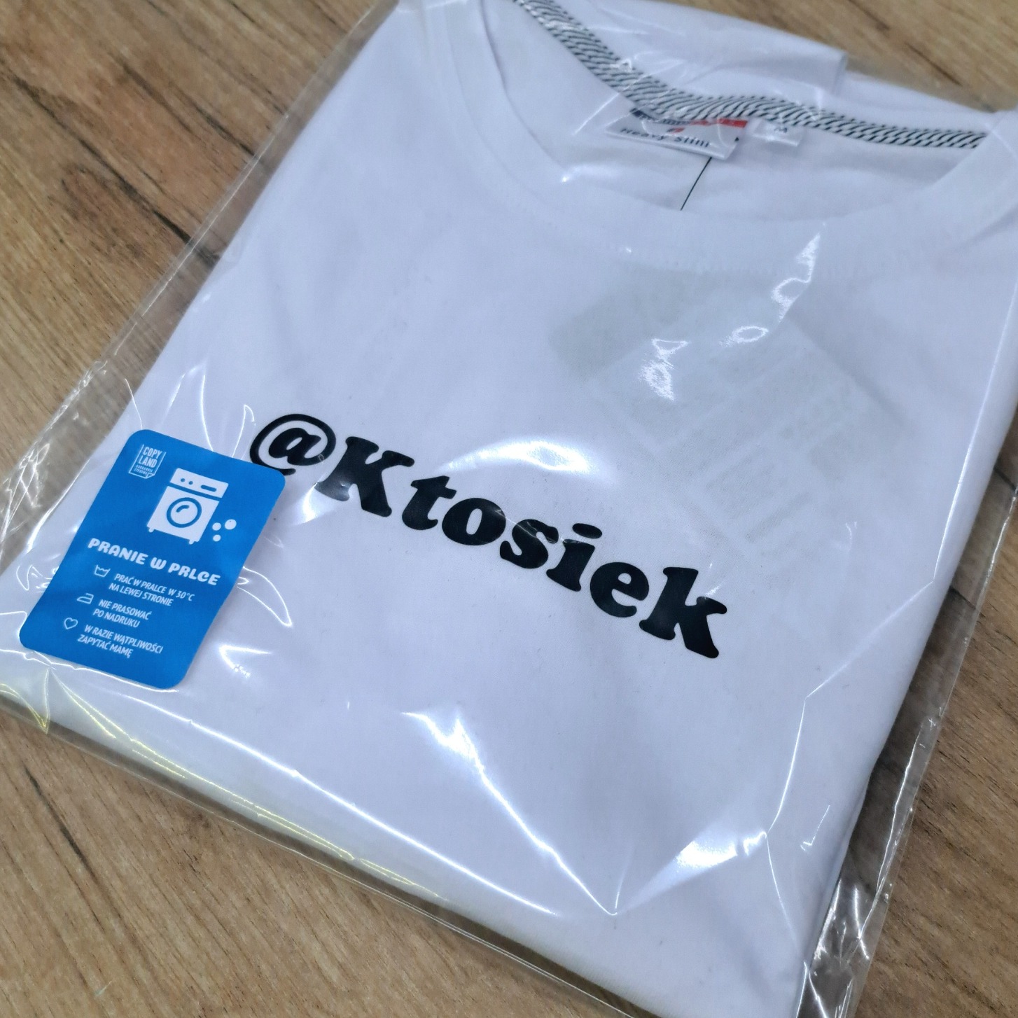 Biała koszulka z czarnym nadrukiem '@Ktosiek' w przezroczystym opakowaniu foliowym, leżąca na drewnianej powierzchni. Widoczna etykieta z instrukcją prania.