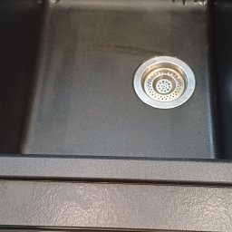 Clean & Care Heroes Marzena Dominik-Kublik - Czysty, czarny zlew granitowy z metalowym odpływem, po sprzątaniu. Widoczne ślady po wodzie i odbicie światła. W tle fragment podłogi z płytek.