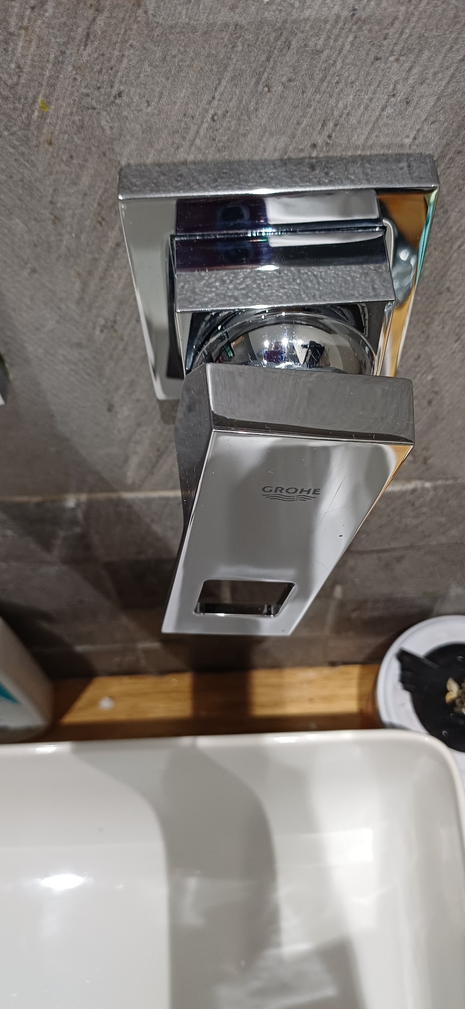 Błyszcząca, chromowana bateria umywalkowa Grohe, zamontowana na szarej, teksturowanej ścianie, z widocznym fragmentem białej umywalki i drewnianego blatu.