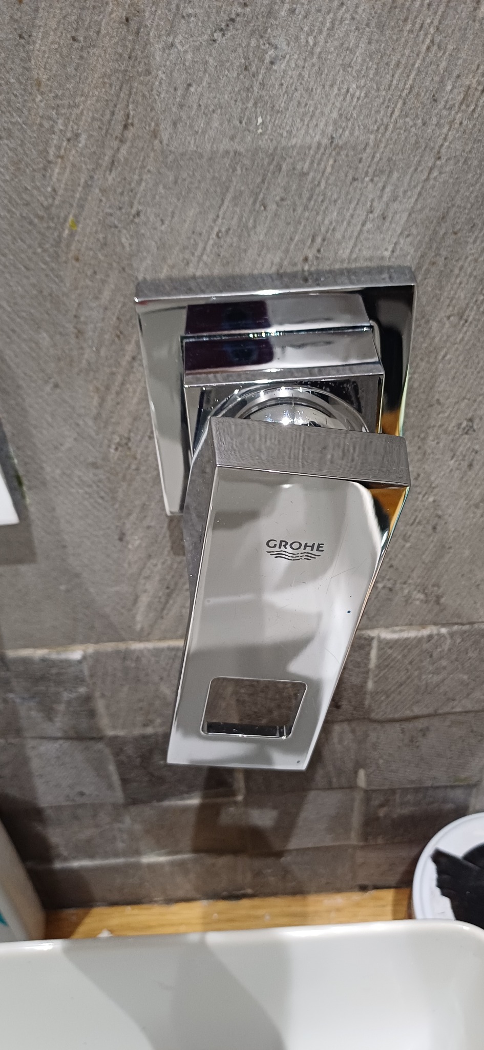 Błyszcząca, chromowana bateria umywalkowa Grohe, zamontowana na szarej, teksturowanej ścianie. Widoczny fragment umywalki i akcesoriów.