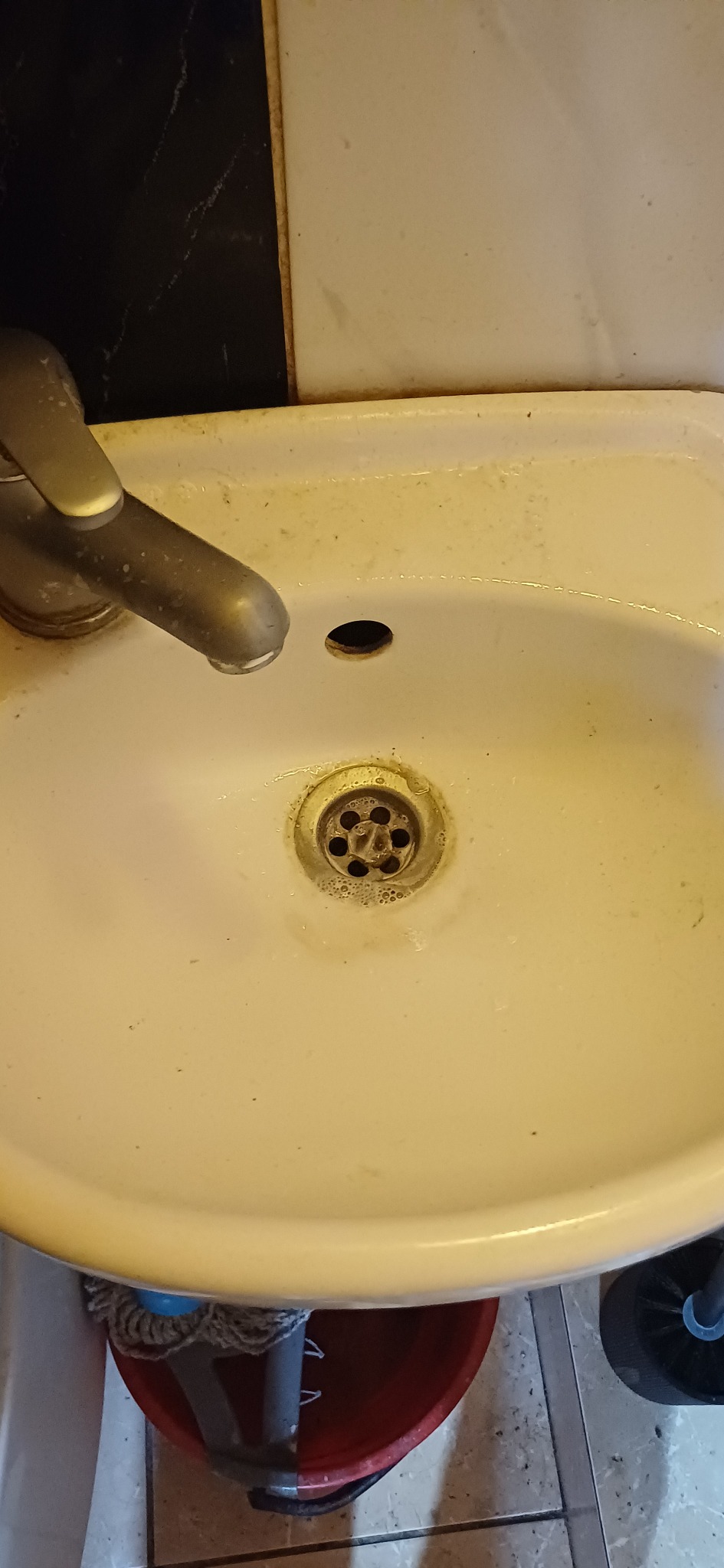 Zanieczyszczona umywalka z widocznym odpływem, kranem i fragmentem mopa w wiadrze. Widoczne ślady użytkowania i zabrudzenia na powierzchni umywalki.