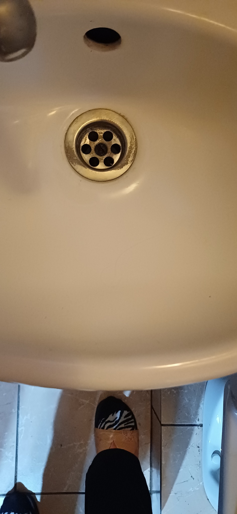 Biała umywalka z odpływem, widok z góry, fragment podłogi z butem w tle. Ujęcie na detale czystości i stanu umywalki.