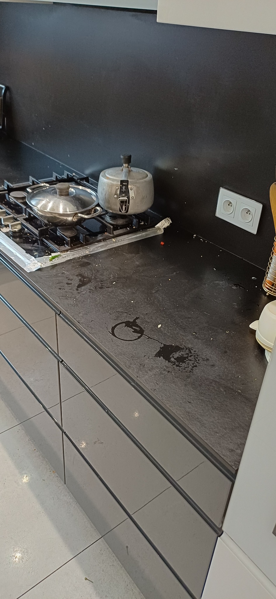 Zabrudzony blat kuchenny z resztkami jedzenia, garnkami na płycie gazowej i plamami po napojach. Widoczne ślady użytkowania i zaniedbania w kuchni.