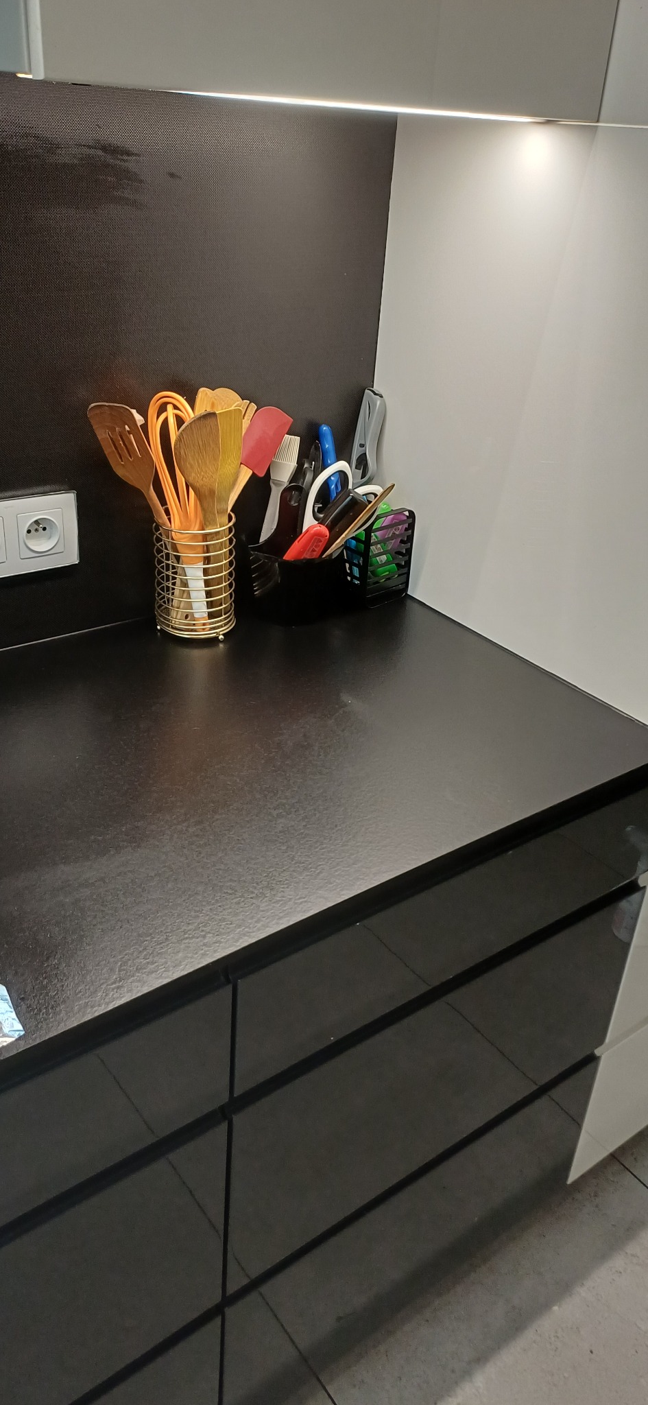 Uporządkowany blat kuchenny z przyborami w pojemnikach: drewniane łyżki, łopatki, pędzle i akcesoria biurowe. Czysta, błyszcząca powierzchnia blatu i minimalistyczny design.