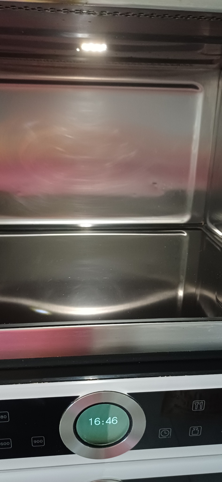 Wnętrze czystego piekarnika z oświetleniem halogenowym, widoczne odbicia na metalowej powierzchni. Panel sterowania z wyświetlaczem czasu na pierwszym planie.