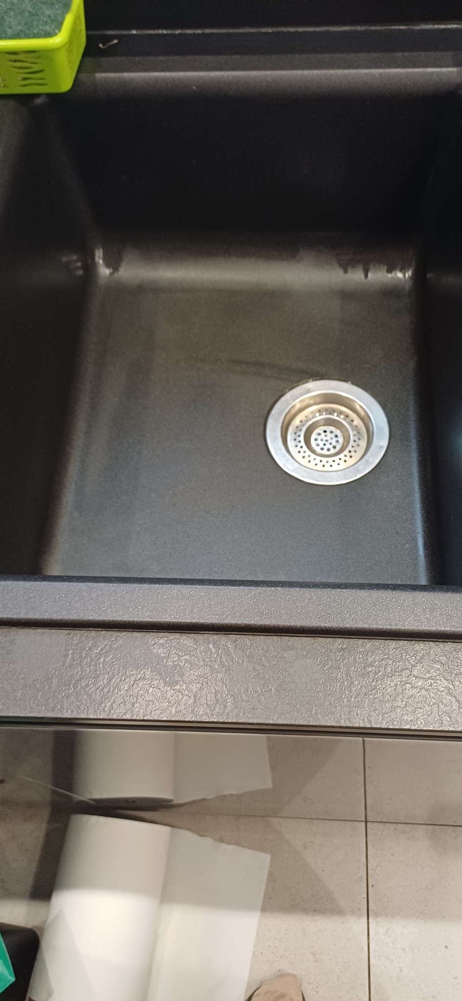 Czysty, czarny zlew granitowy z metalowym odpływem, po sprzątaniu. Widoczne ślady po wodzie i odbicie światła. W tle fragment podłogi z płytek.