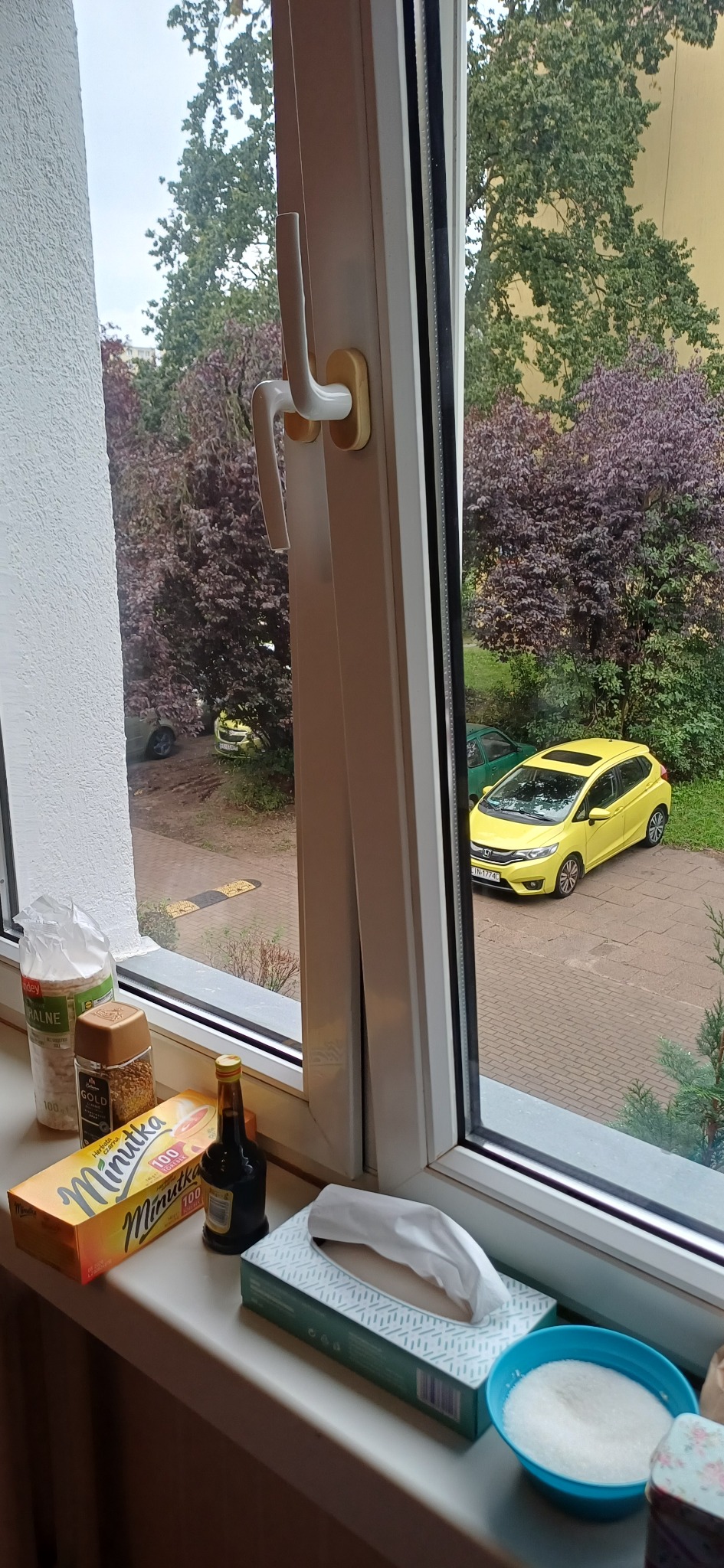 Widok z okna na żółty samochód na parkingu, z bliska widoczne produkty spożywcze i higieniczne na parapecie. Biała rama okienna z uchwytem.