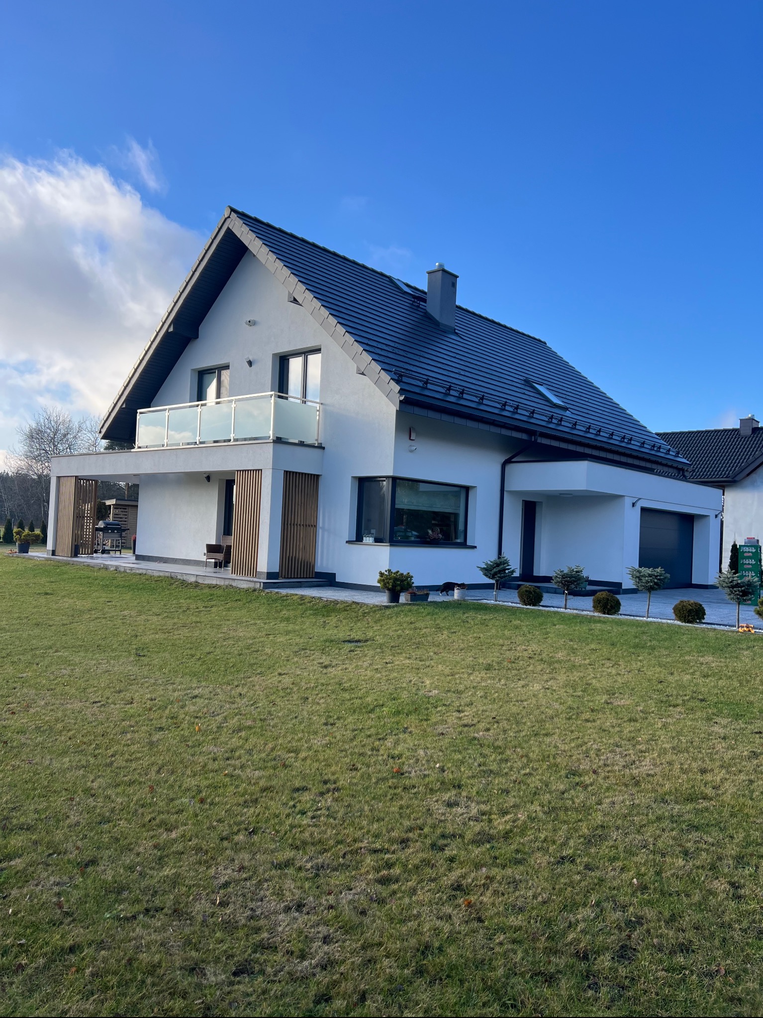 Nowoczesny dom jednorodzinny z białymi ścianami i ciemną dachówką, widok z trawnika w słoneczny dzień. Balustrada balkonowa ze szkła. Elementy dekoracyjne z drewna.