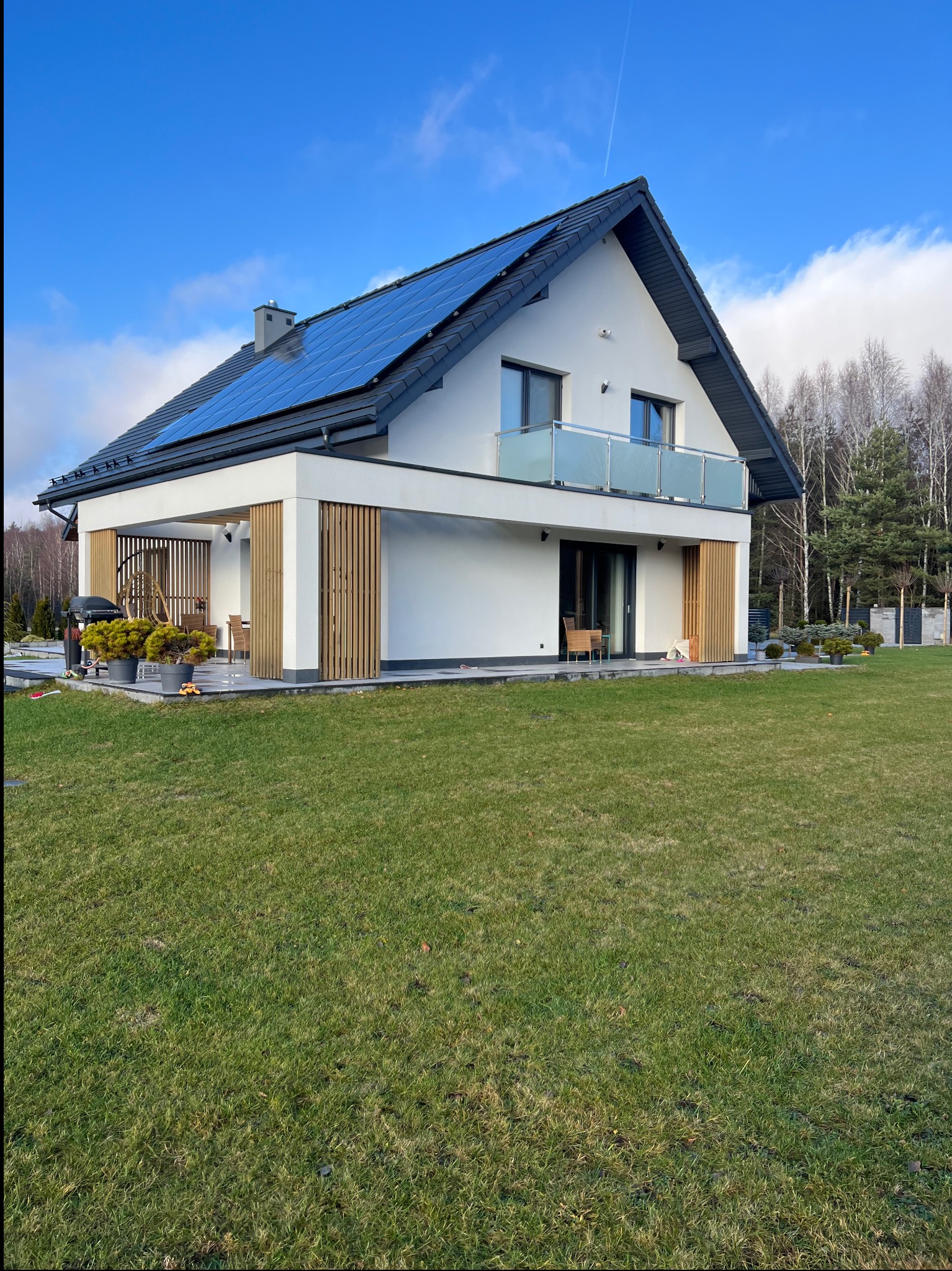 Nowoczesny dom jednorodzinny z białą elewacją, panelami słonecznymi na dachu i drewnianymi elementami dekoracyjnymi. Widok z zadbanego trawnika w słoneczny dzień.