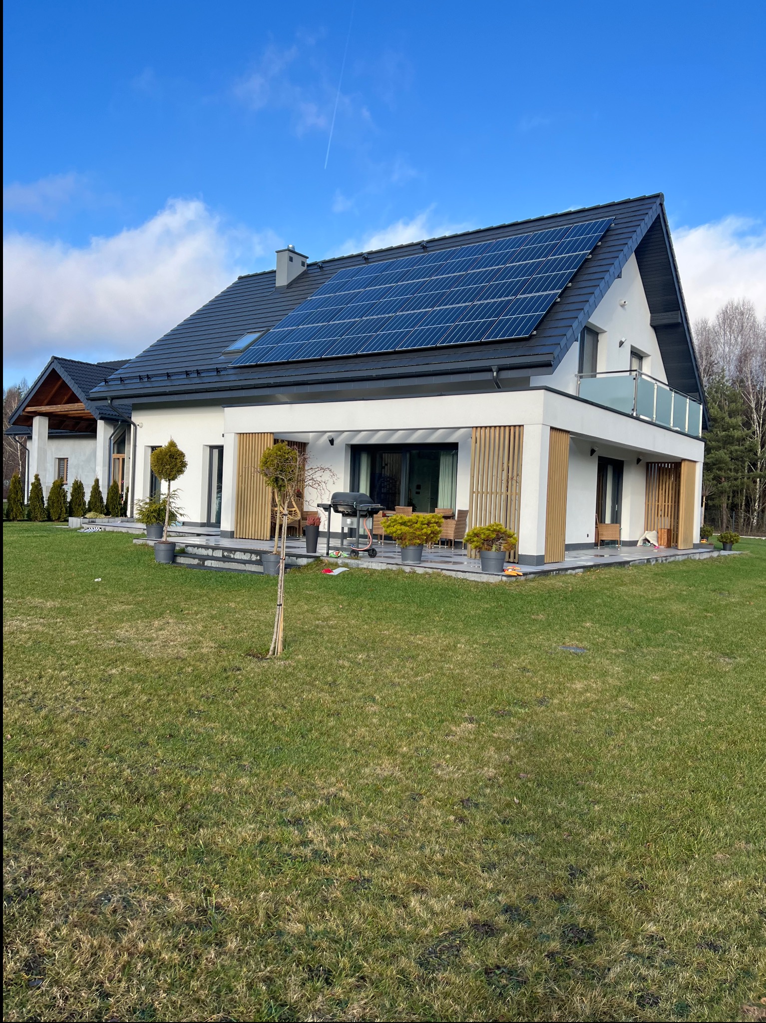 Nowoczesny dom jednorodzinny z białą elewacją, panelami słonecznymi na dachu i drewnianymi elementami dekoracyjnymi na tarasie. Widok z ogrodu w słoneczny dzień.