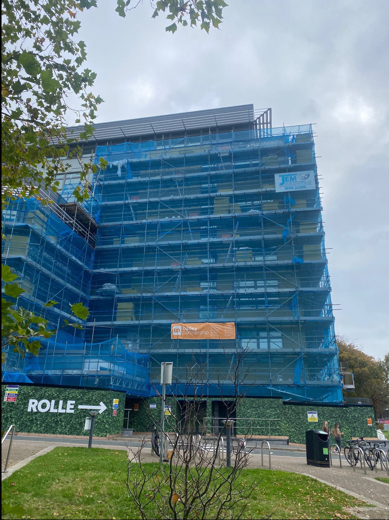 Wielopiętrowy budynek w trakcie prac elewacyjnych, osłonięty rusztowaniami i siatką ochronną. Widoczne logo firmy na rusztowaniu i tymczasowe ogrodzenie terenu budowy.