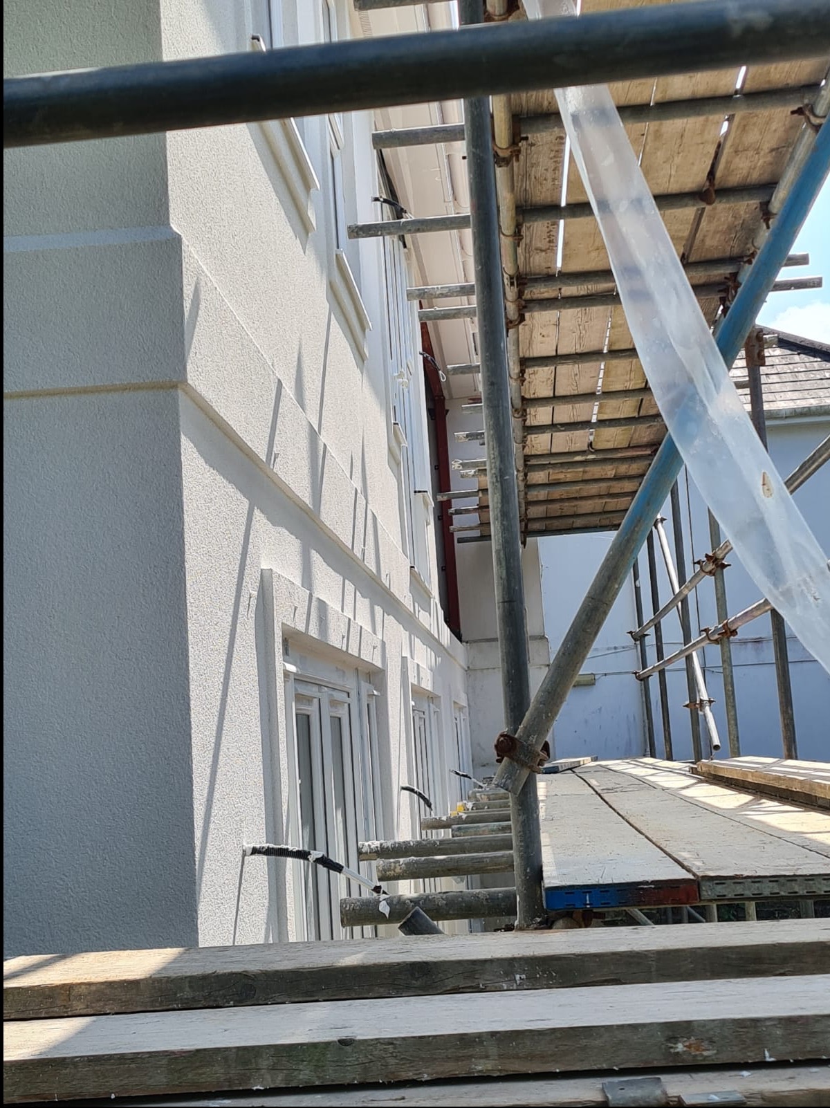 Świeżo otynkowana, jasna elewacja domu z widocznymi detalami architektonicznymi, częściowo zasłonięta przez rusztowanie. Słoneczny dzień.