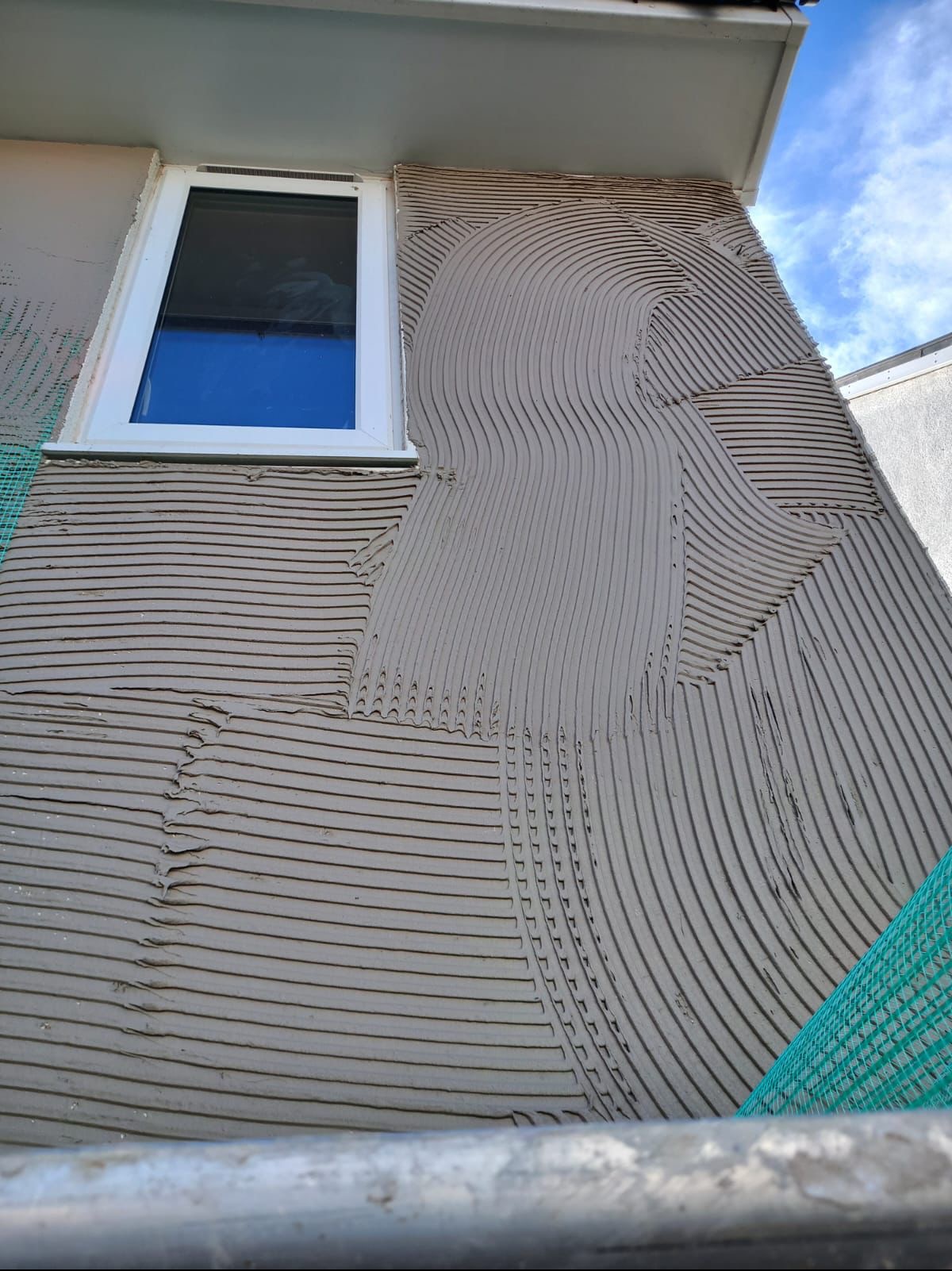 Ściana budynku z nałożoną warstwą kleju pod ocieplenie, widoczna siatka elewacyjna i okno. Charakterystyczny wzór kleju w łuki i proste linie.