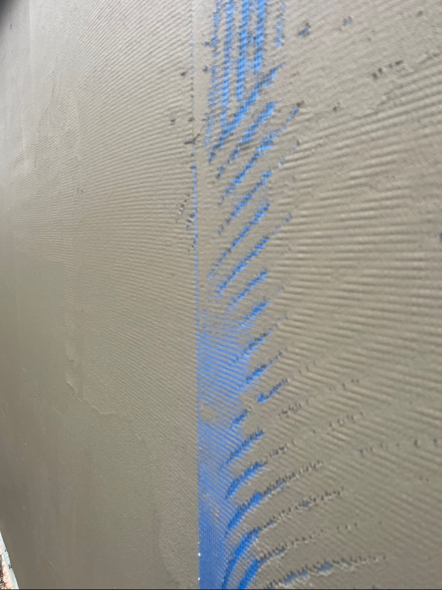 Świeżo nałożona warstwa kleju na elewacji z widoczną siatką zbrojącą, przygotowana do dalszych prac wykończeniowych. Charakterystyczna faktura kleju.